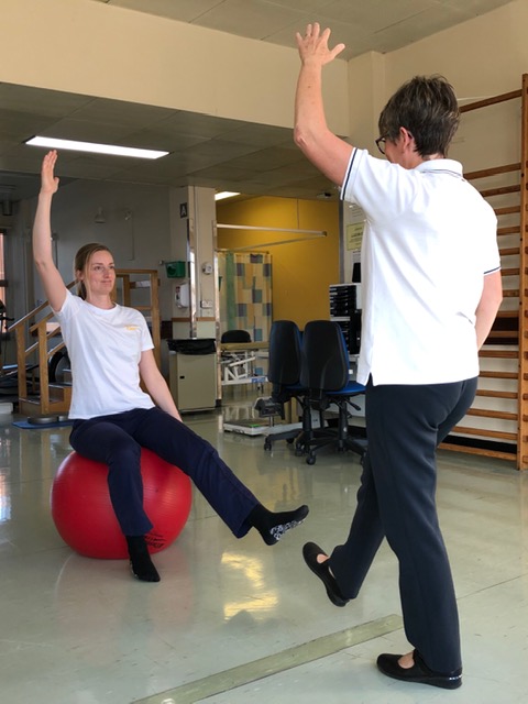 An exercise rehab session with a physiotherapist showing a patient on a ball how to perform a balance exercise.