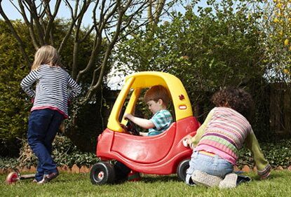 Photograph of children playing outdoors. Little boy sitting in a brightly coloured plastic car.