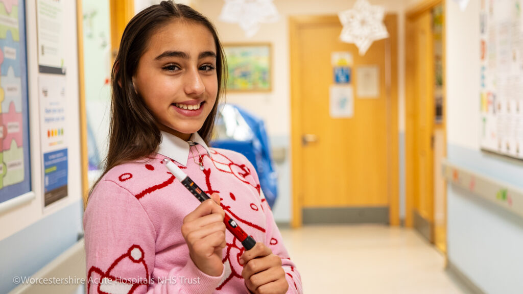 A picture of girl smiling on a children's ward setting in a hospital, holding an insulin pen.