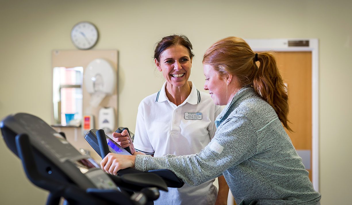 A physiotherapist smiling at a patient who is using a piece of sports equipment in a hospital setting. A door, clock, and noticeboard are visible in the background.