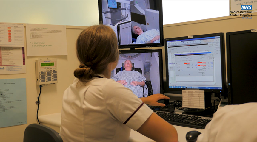 An image of a radiographer sat at their desk whilst the scan is progressing. They are watching the patient on a live screen and have their computers and scanner screens open 