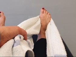 Feet on bed toes up, towel rolled and wrapped around base of one foot towel ends in both hands