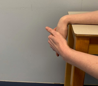 An image of a forearm resting on a table the wrist on the edge and the hand dangling over the side. The other hand is applying gentle pressure to push the hand inwards