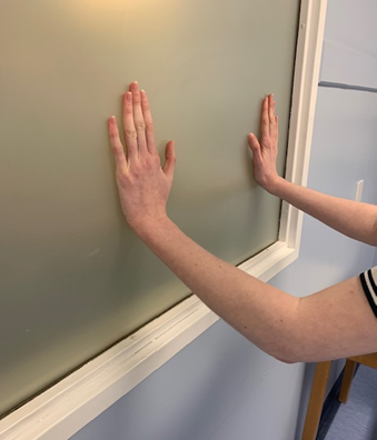 An image of an individual doing a wall press-up. They are stood facing a wall with arms straight and hands pressing into the wall