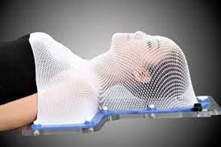 Image of a person lay flat on back on bed having a thermoplastic mesh head mould made. Their head, chest and shoulders are covered with the white mesh which is moulding to their facial features