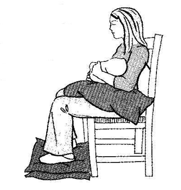 An individual sat in a chair with a pillow on lap supporting a child in their arms and pillows stacked under their feet so they can rest them securely