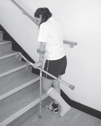 An individual preparing to walk up stairs using one crutch and stair rail but carrying the other crutch whilst holding the usable crutches handle