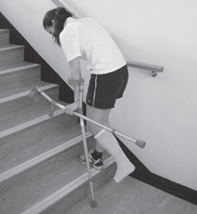 An individual walking up stairs using one crutch and stair rail but carrying the other crutch whilst holding the usable crutches handle  their weight is on the crutch and stair rail 