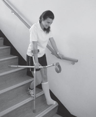 An individual preparing to walk down a step using one crutch and stair rail but carrying the other crutch whilst holding the usable crutches handle