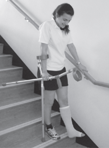 An individual preparing to walk down another step using one crutch and stair rail but carrying the other crutch whilst holding the usable crutches handle