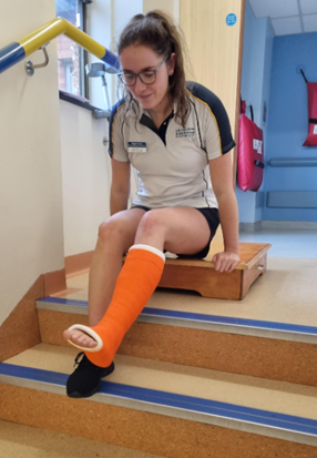 An individual in a leg plaster cast is preparing to stand having reached the top of the stairs on their bum. They are  using both hands to push themselves onto a small stool (away from the top of the stairs) to make this transition easier