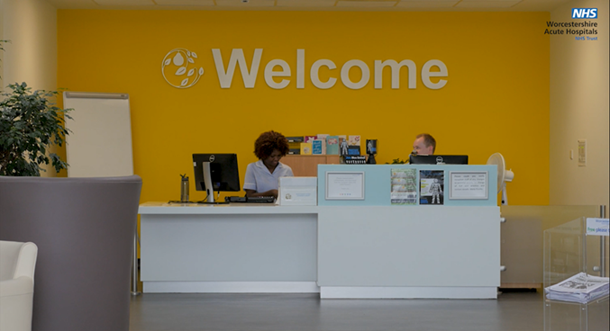 A photographic image of the reception area of Worcestershire Oncology Centre at Worcester Royal Hospital showing the welcome desk, reception staff and the orange wall with Welcome signage