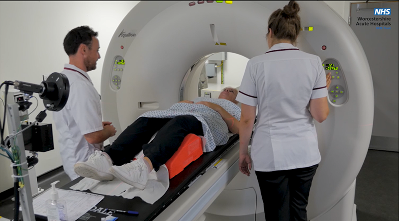 An image of a patient being guided into one of the scanners accompanied by two Radiology staff. The patient is lay on their back on the bed with knees supported. They are wearing a gown. Everyone looks calm and happy