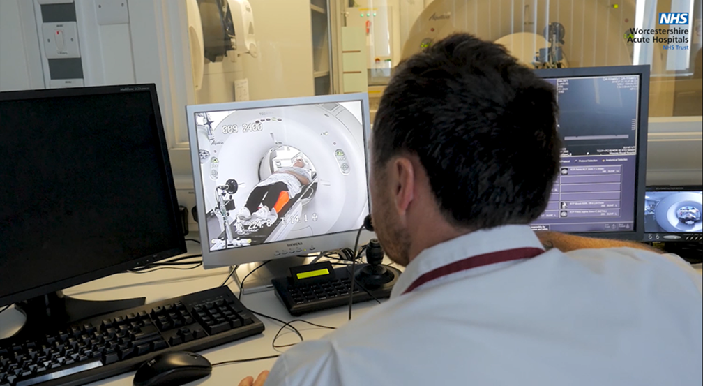 An image of a radiographer sat at their desk whilst the scan is progressing. They are watching the patient on a live screen and have their computers and scanner screens open 