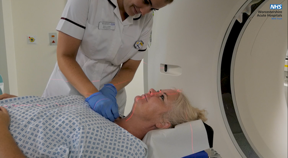 An image of a patient having a small tattoo applied whilst they lay on the scanner bed. They are wearing a hospital gown and are smiling