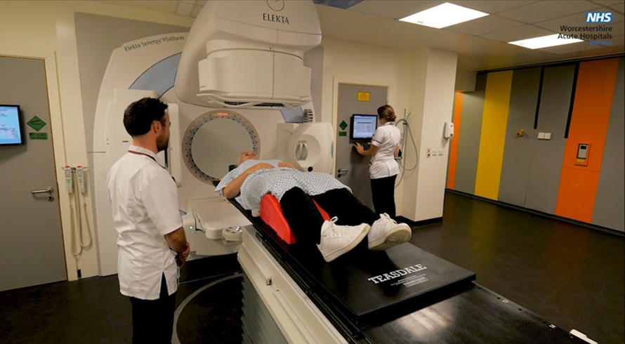 An image of a patient being positioned for a scan. Patient is lying on their back on a bed with a radiologist to there side. A second radiologist is preparing the scan on the display screen.