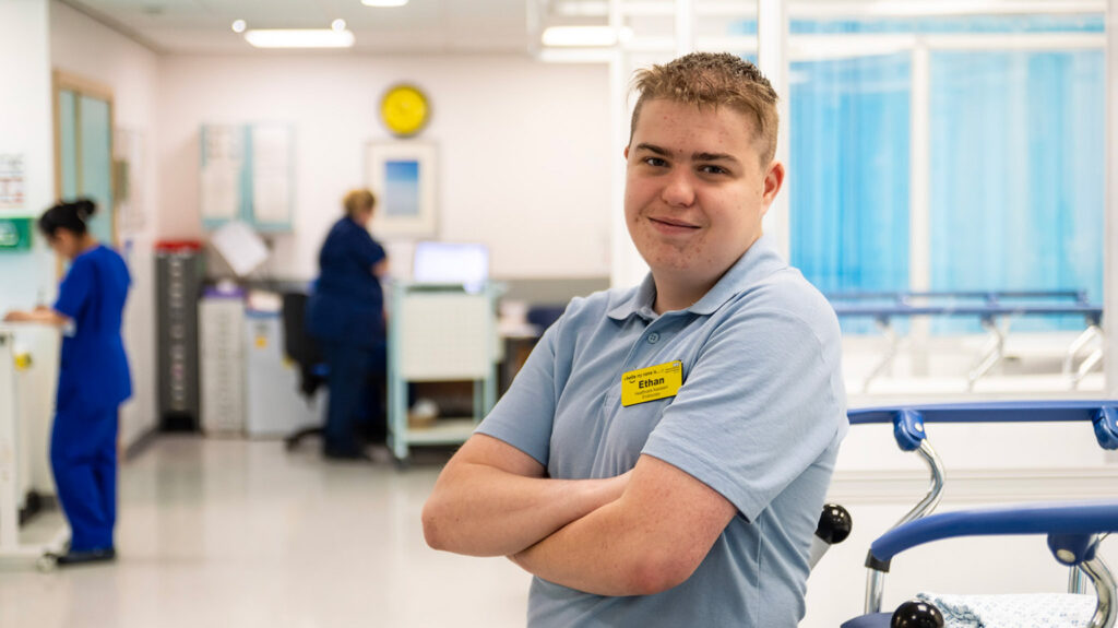Photograph of Ethan, smiling to camera on a hospital ward. Background shows staff member and equipment blurred.