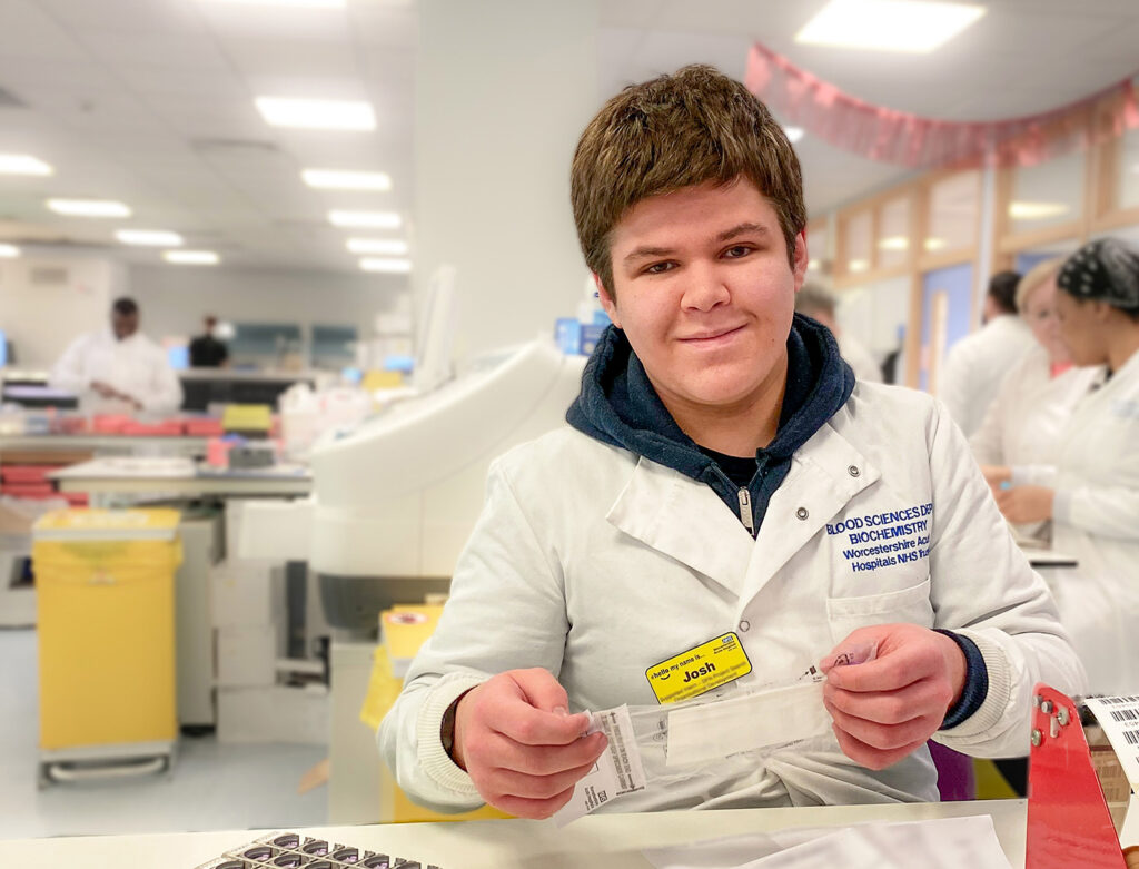Photograph of Josh, young intern in the laboratory. Holding paperwork and smiling at camera. Behind him a busy laboratory backdrop.