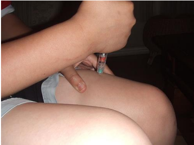 Picture of a person pinching the left thigh with left hand and in the right hand is a needle that is punctured through the skin of the pinched left thigh