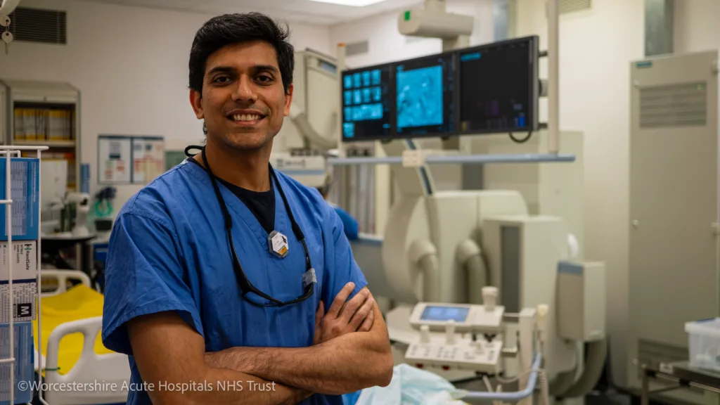 Dr Rahul Chivate wearing blue scrubs stood smiling at camera with his arms folded in the Interventional radiology suite.