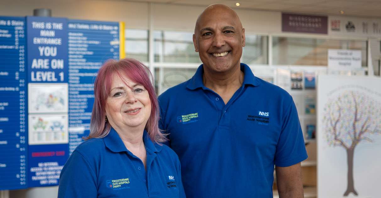 Photo of two volunteers standing in the foyer of Worcestershire Royal Hospital. Smiling at camera. Volunteers wearing the volunteer t-shirt.