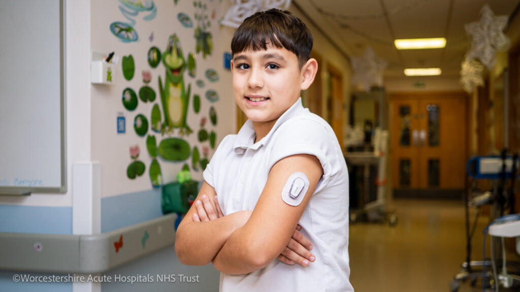 Boy stood on a children's ward setting in a hospital, with a glucose monitoring patch on his arm looking at the camera and smiling.