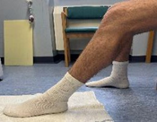 Image of person sat on stool with feet in socks flat on floor and one foot on a towel. Leg with foot on towel is extended in front as if towel has been pushed along floor by foot