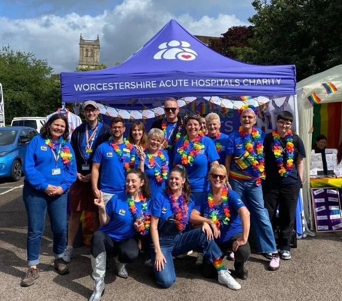 Photograph of the LGBQT+ team at an event. The team are standing underneath a blue pop up tent with the branding Worcestershire Acute Hospitals Charity.