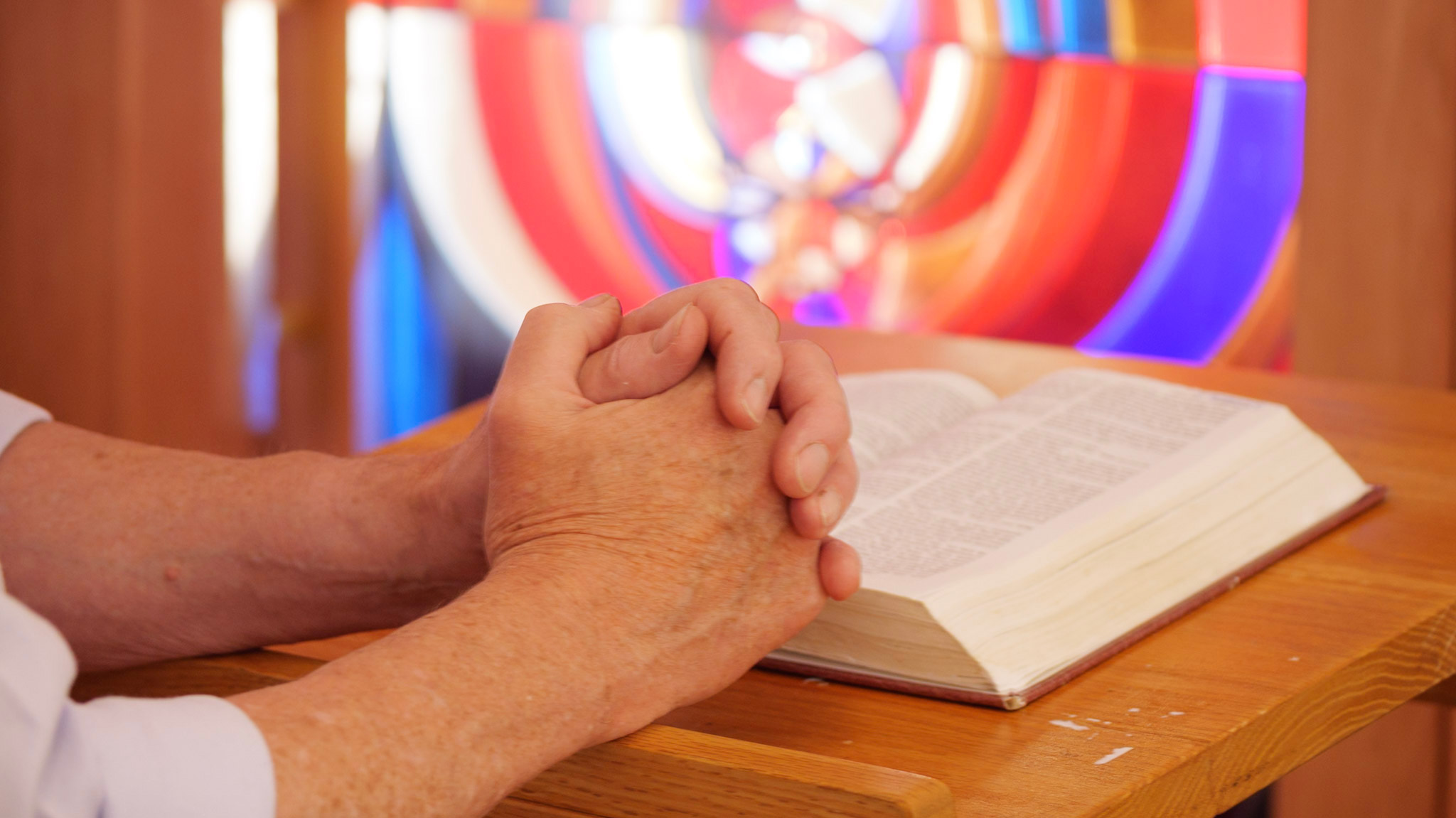 Hands perched together on a bible. In the background a beautiful coloured window is reflecting colours.