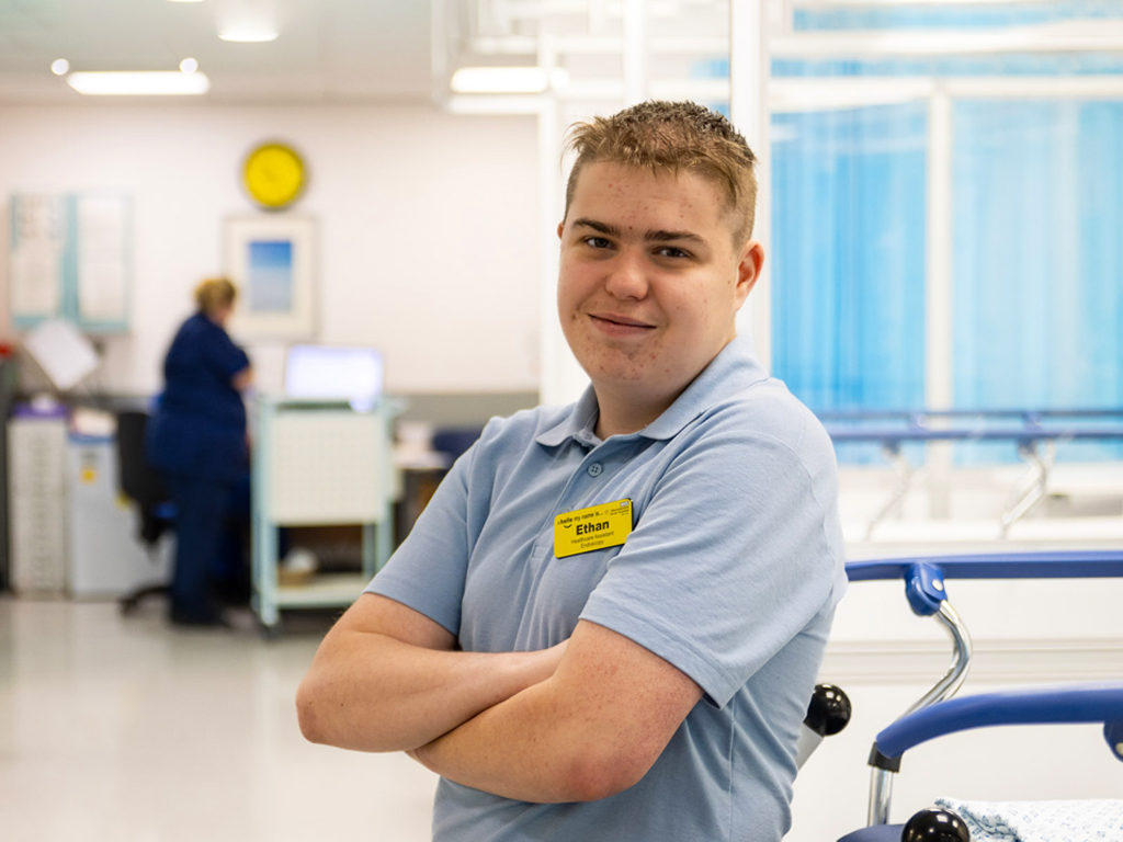 Ethan standing and smiling on a hospital ward, with staff members visible in the background slightly out of focus.