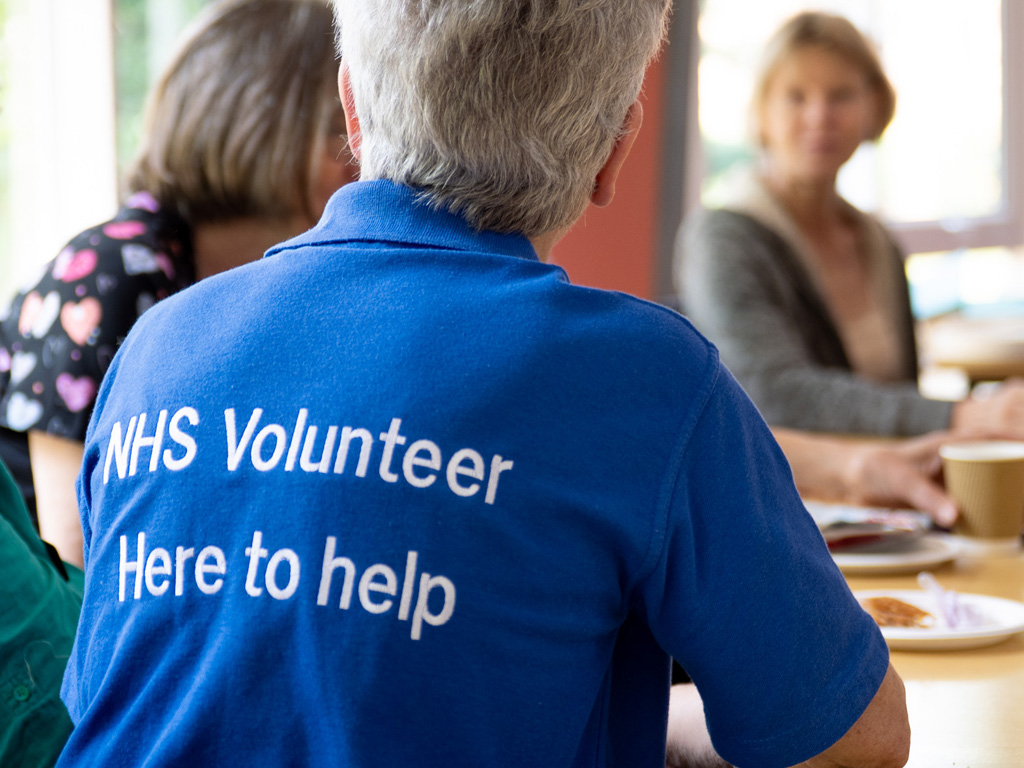 NHS volunteer sitting at a table, smiling and talking to people. Her T-shirt reads “NHS Volunteer – We’re here to help.”