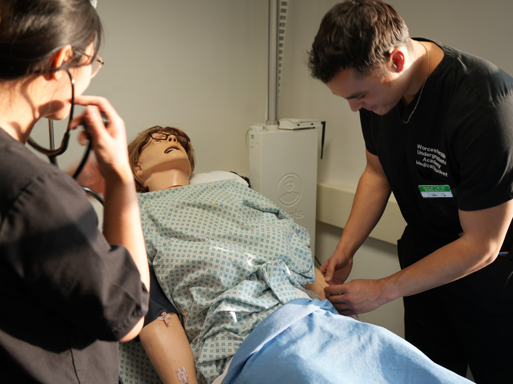 Two medical students in a simulation room at the Charles Hastings Education Centre, leaning over a medical mannequin while preparing for a clinical examination.