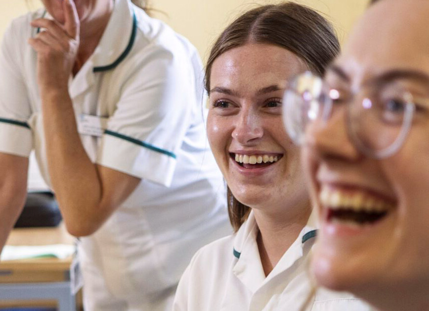 Close-up candid photo of Allied Health Professionals smiling and laughing. Central focus on a young woman smiling. Head and shoulders visible in a hospital setting.