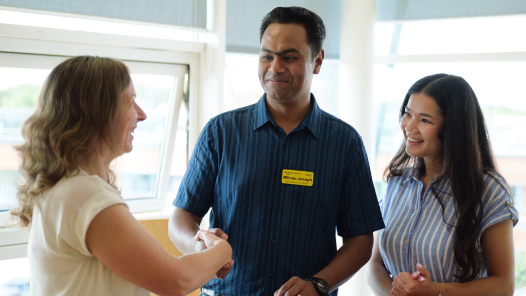 Three people photographed in room. One of the people is shaking the hand of the other. Other person is looking on and smiling. Background of room shows brightly lit windows.
