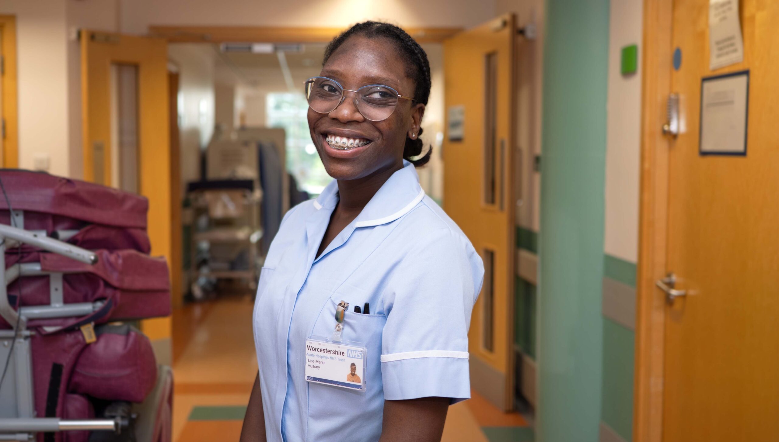 Photograph of Healthcare Support worker smiling at work. The portrait photo shows her standing on a hospital ward head and shoulders. In the background you can make out the equipment on the ward