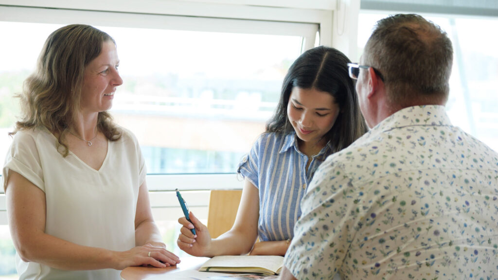 Photograph of three people in a meeting. Picture has captured a moment of the meeting where the three people are Smiling, chatting. Office background looks out on large windows where light is reflecting into the room.
