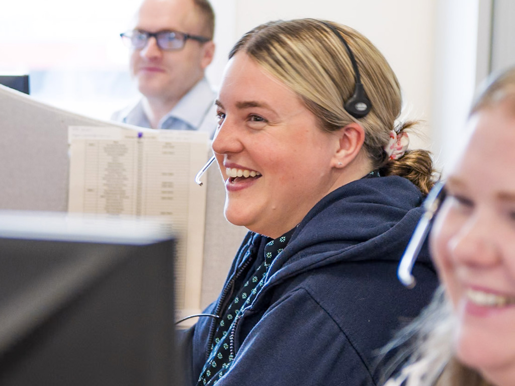 Smiling woman wearing a headset, sitting at a desk in an office with colleagues blurred in the background.