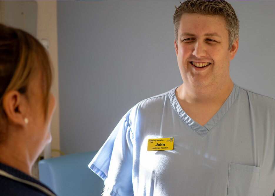 Photograph of Healthcare Assistant smiling and talking to nurse on hospital ward.