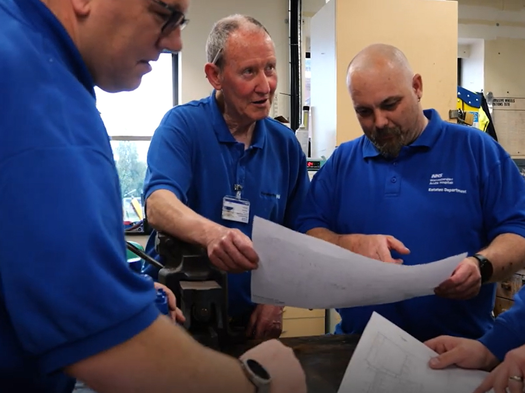 Photograph of three men from Estates and facilities in busy workshop. Holding paperwork and plans of building.