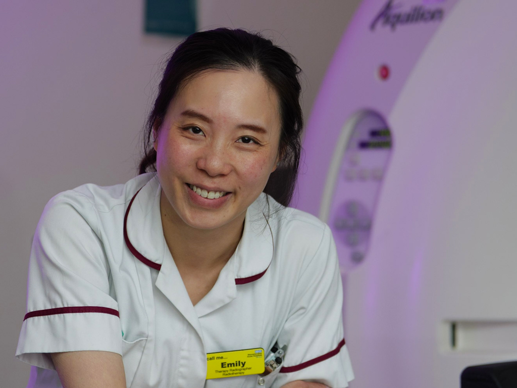 Smiling therapy radiographer in uniform, head and shoulders visible in front of a CT scanner.