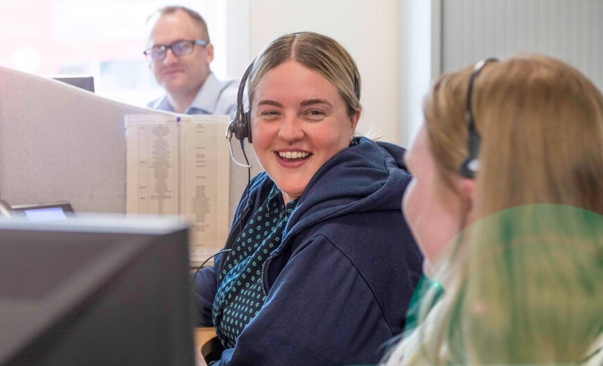 Smiling photo of administration member of staff in hospital. Headset piece over head. Smiling and talking to work colleagues in office.