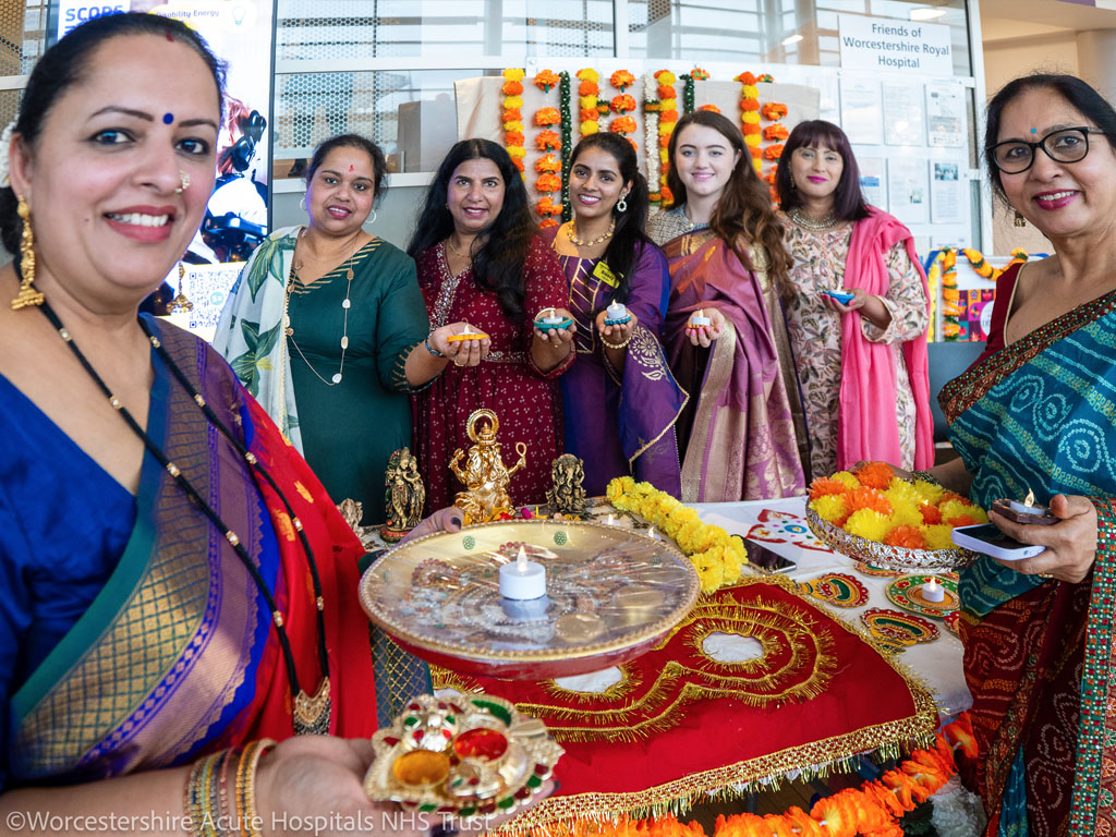 A photograph of Hospital staff dressed in colourful sarees in the foyer of Worcestershire Royal Hospital celebrating Diwali. The scene includes a vibrant Rangoli display symbolising positivity, a table with sweets representing joy and good fortune, informative leaflets about Diwali’s cultural significance.
