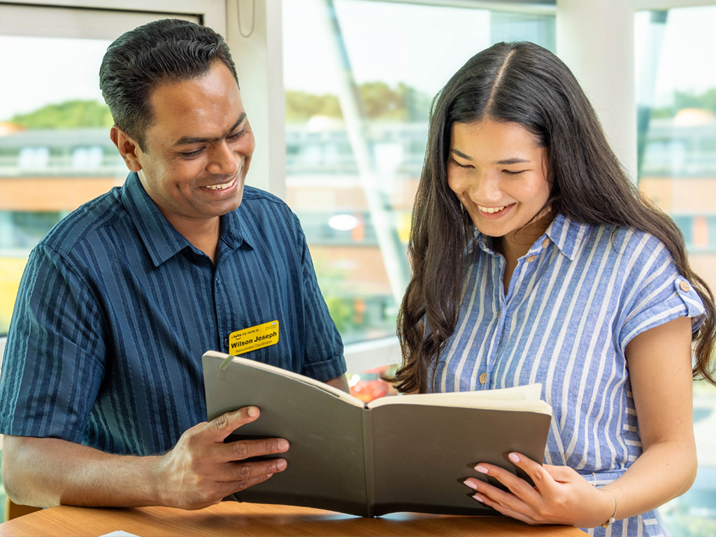 Two members of the Recruitment team sitting in an office, smiling as they look over paperwork, with brightly lit windows and slightly blurred hospital buildings in the background.