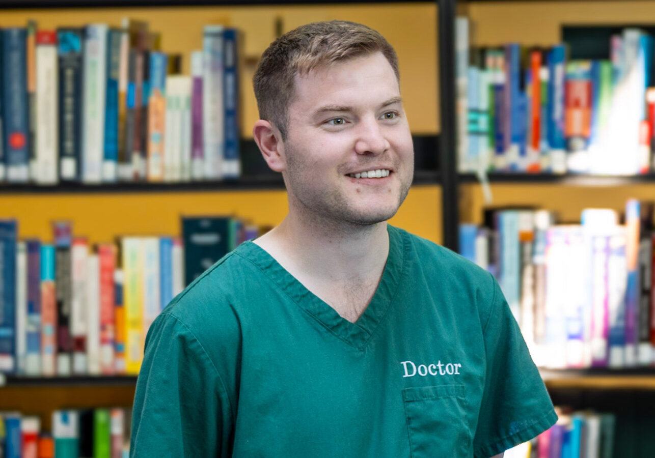 Photograph of a young male doctor. Smiling. This is a portrait photograph. Behind him are lots of books on a book shelf.