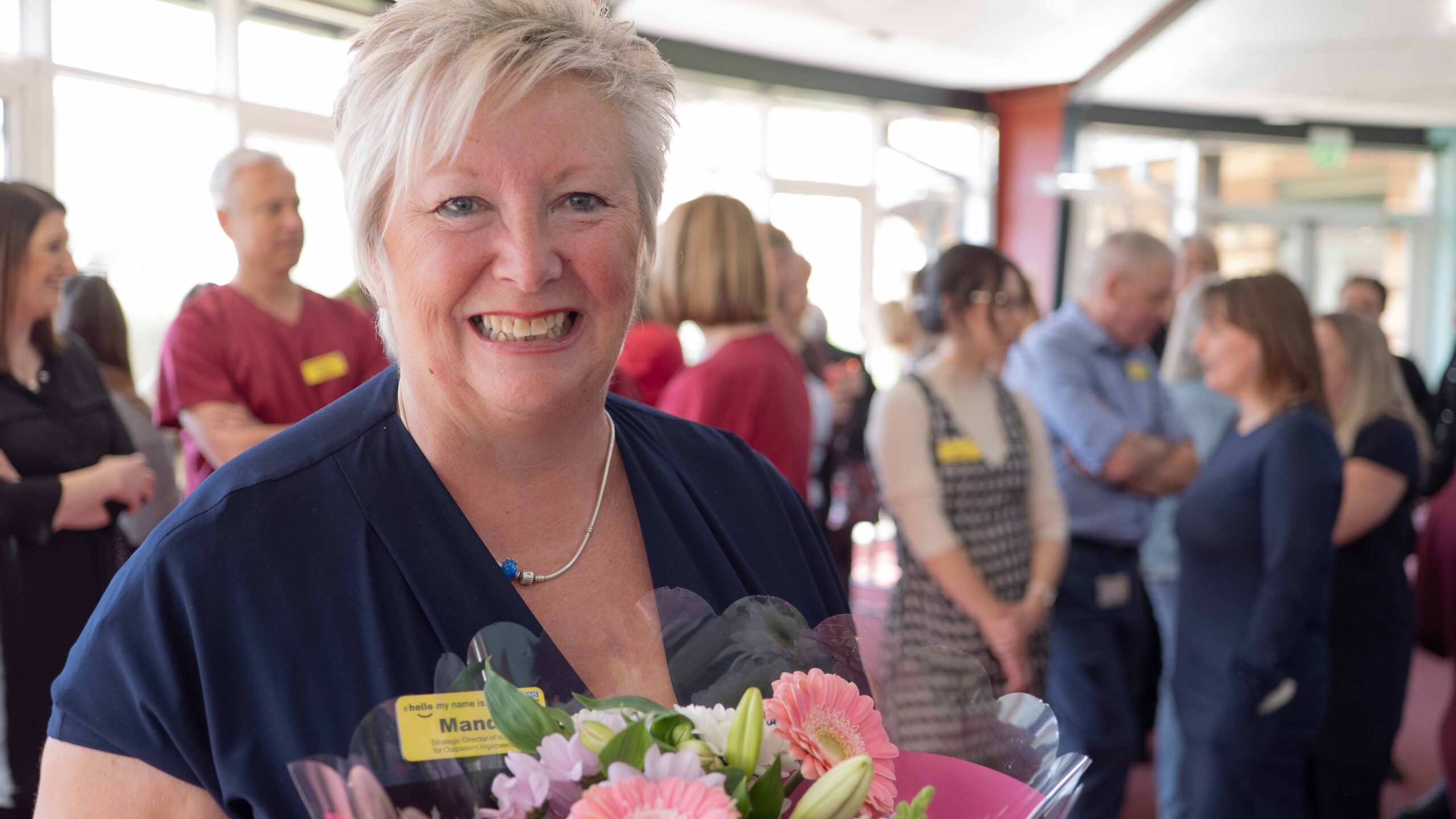 Smiling woman receiving a colourful bouquet of flowers, with a softly blurred background showing people engaged in conversation