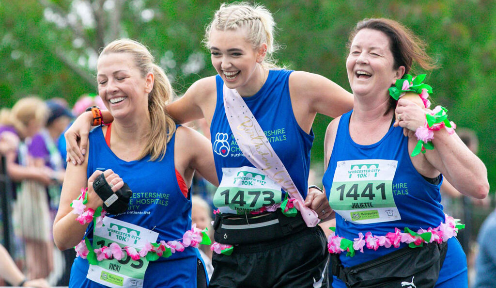 Picture of three runners at Worcester City Run. Smiling and laughing as they run. Arms are linked as they run. Middle runner is wearing a birthday sash.