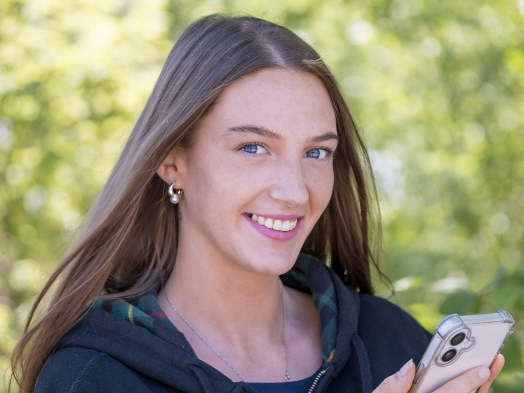 Young woman smiling at the camera, holding a mobile phone. She is outdoors on a sunny day, dappled leaves and bushes blurred in the background.
