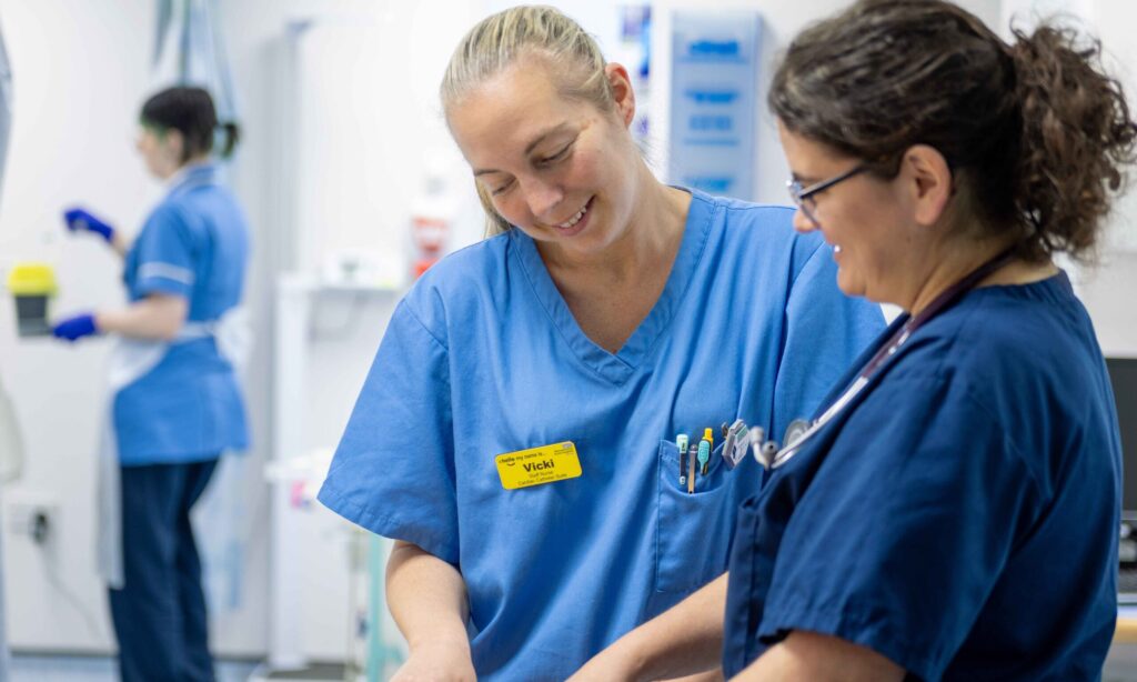 Two healthcare workers in blue scrubs stand together in a clinical setting, smiling and looking down at something they are working on. One has a yellow name badge and several pens in her pocket. Another staff member in blue scrubs works in the background.