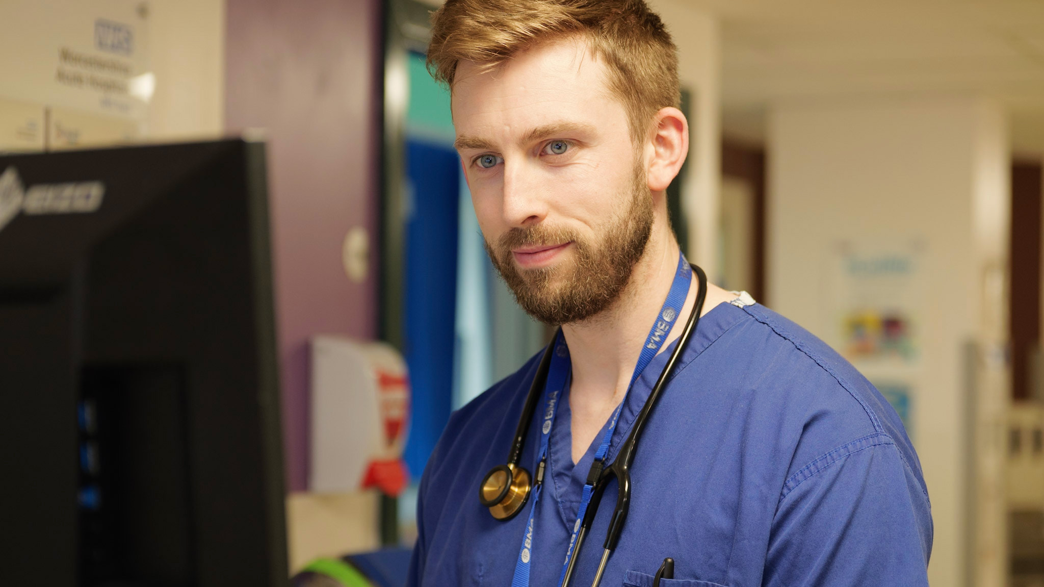 Portrait photo of a doctor on a ward. He is looking at a computer screen. This is a head and shoulder photograph.