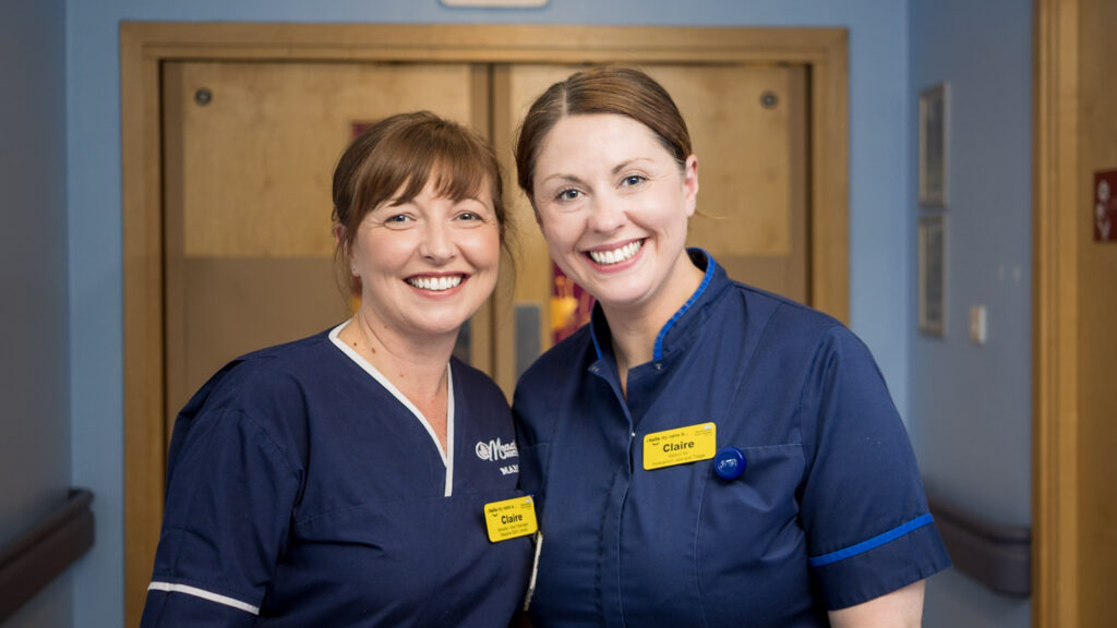 Two midwives in uniform stood smiling at the camera in a hospital setting